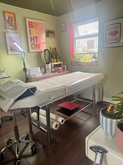 Treatment room: white couch, ring lamp, sage-green walls, "BRIGHTON" art prints, plants, sunlight